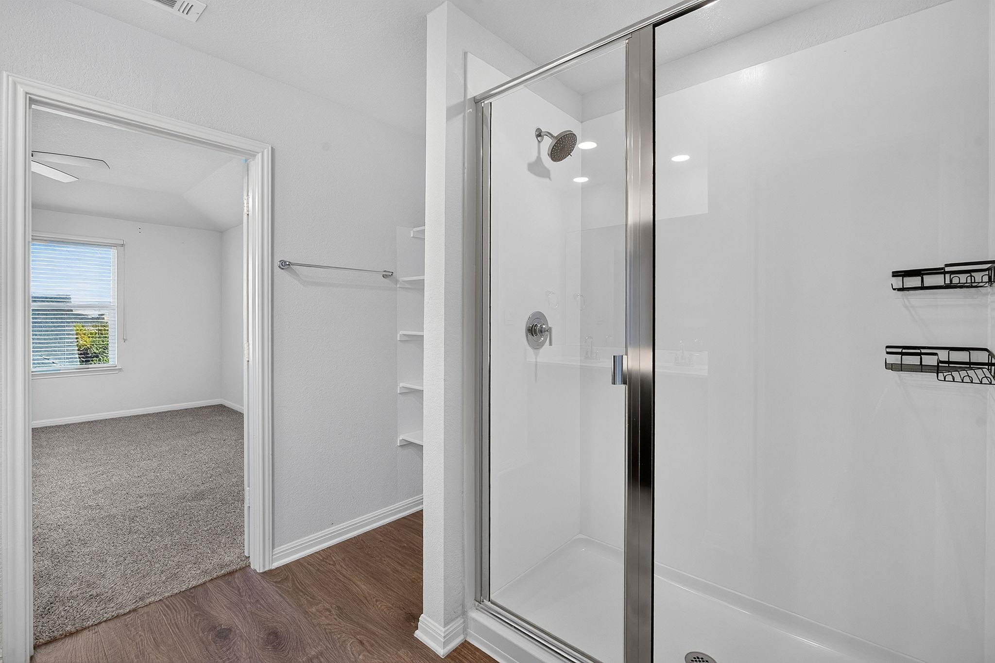 A glass-enclosed shower stands adjacent to wood-look flooring leading into a carpeted bedroom.