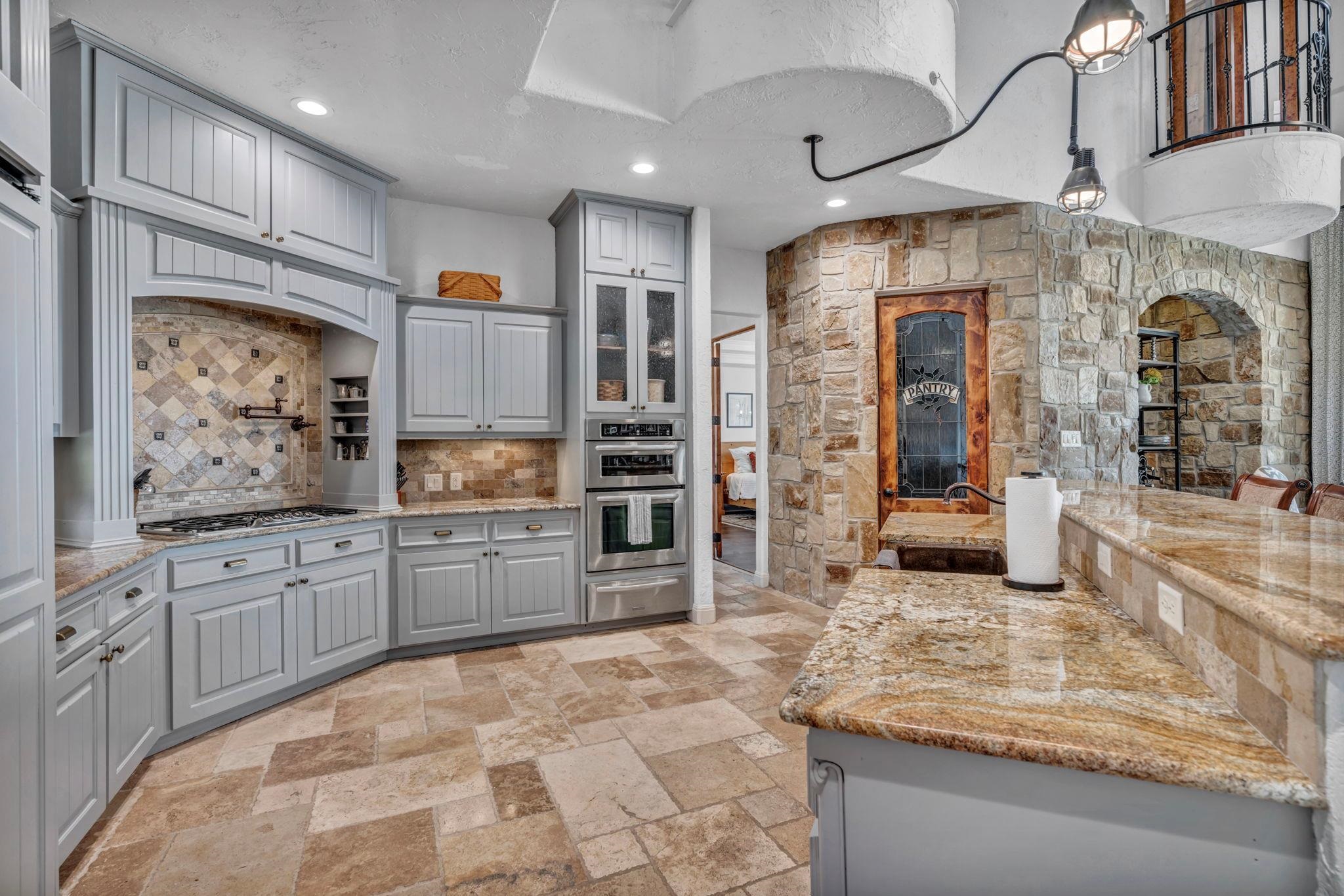 Featuring gray glass insert cabinets, light stone counters, pot filler, double ovens, gas top